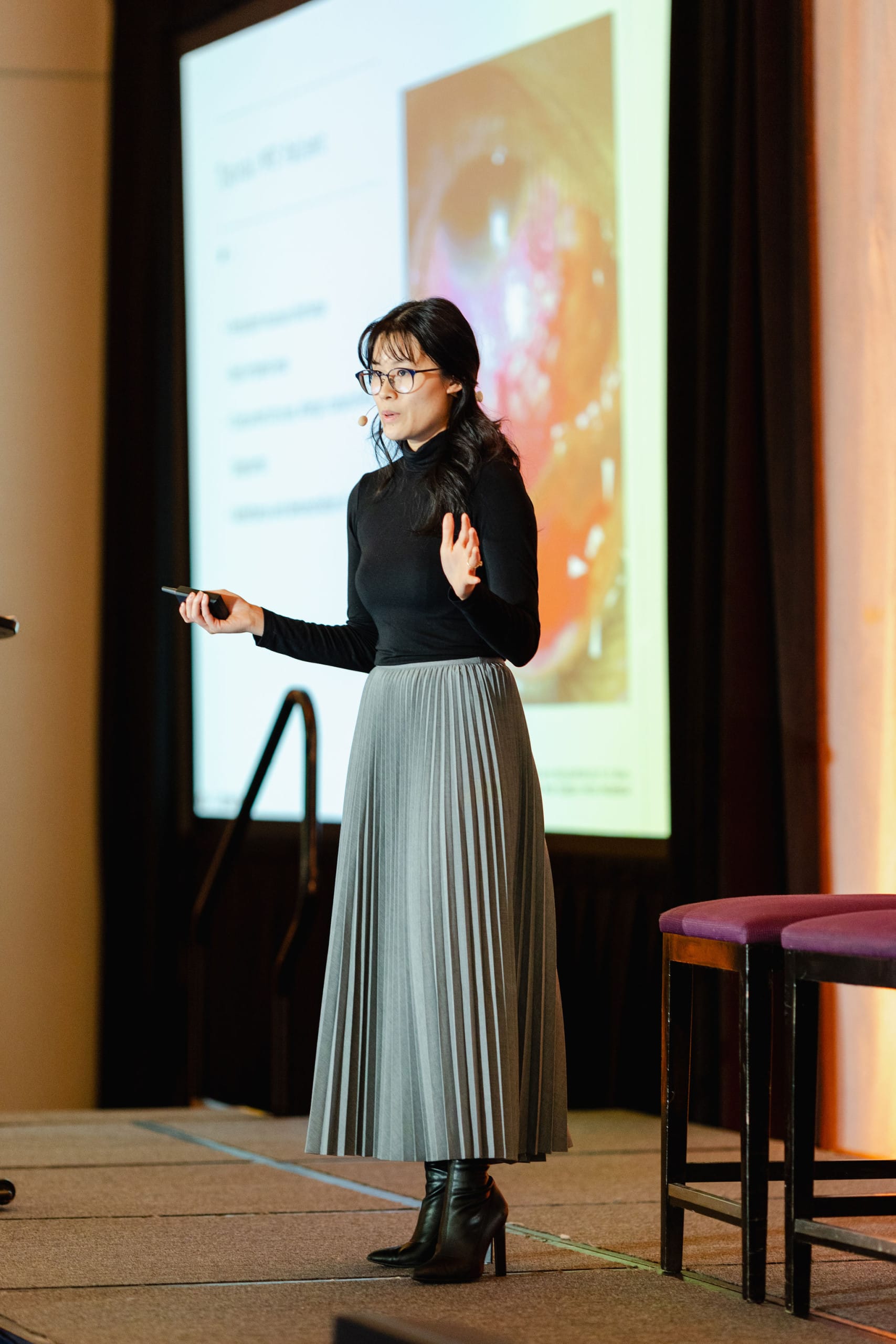 A woman in a black top and gray skirt delivers a presentation on stage, captured through expert event photography. She wears a microphone headset, with a slideshow projected in the background.