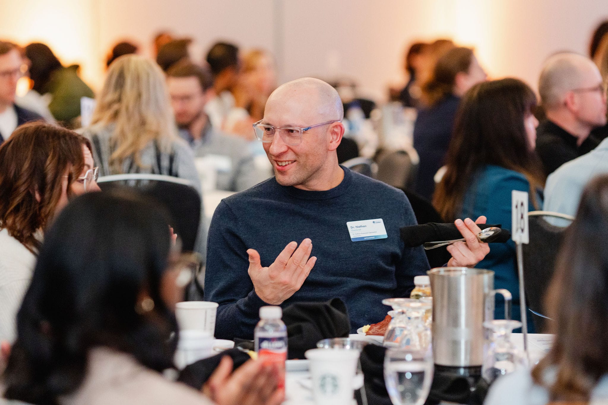 A bald man wearing glasses talks animatedly at a table in a conference setting, captured brilliantly through event photography, surrounded by engaged participants.