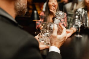 A person holds an intricately designed glass bottle at a table. Three people sit in the background, with wine glasses beside them.