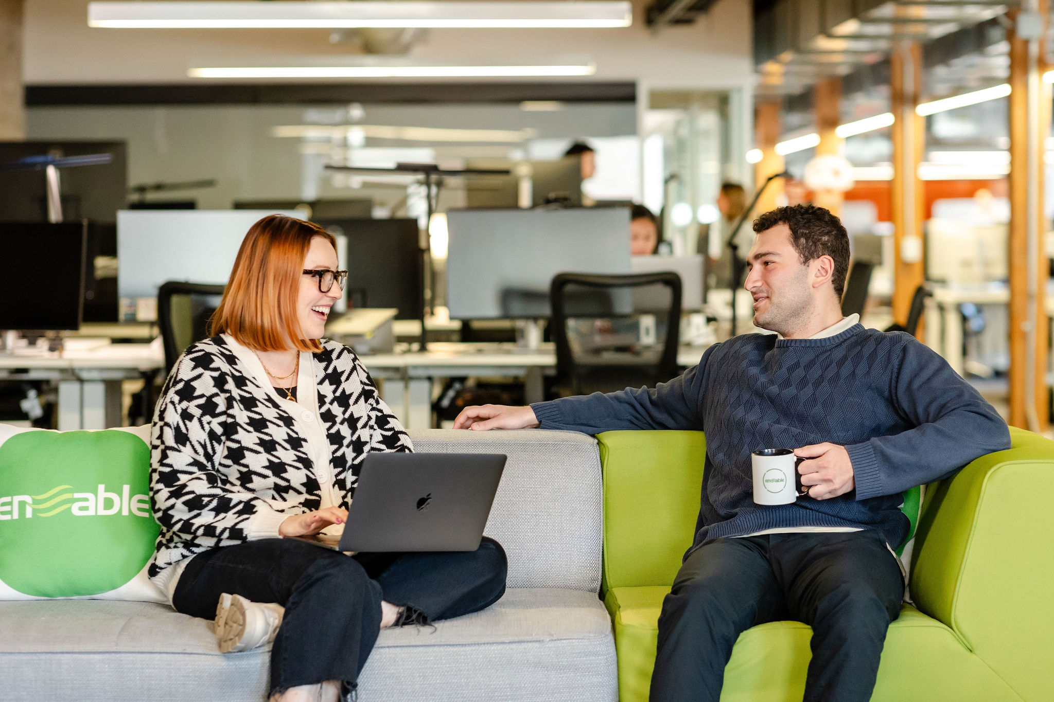 Two people sit on a brightly colored couch in an office environment, engaged in conversation. One has a laptop, the other holds a mug, capturing a candid moment perfect for corporate photography.
