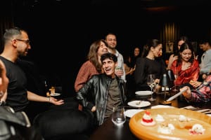 A group of people laughing and talking at a dimly lit dining setting with empty plates and a dessert platter on the table.