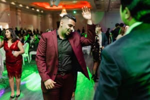 A man in a burgundy suit dances with a raised hand at a party, surrounded by people under colorful lights.