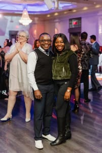 A man and woman pose together, smiling, at a party with other guests in the background.