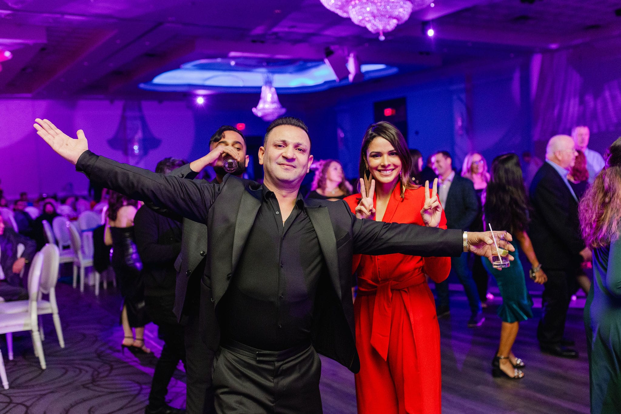 A group of people at a lively event, with one person in front gesturing with open arms, and another making a peace sign. Others are mingling in the background.