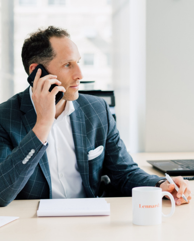 A man in a suit, captured through corporate photography, is on his phone while sitting at a desk equipped with a notepad, pen, and coffee mug.