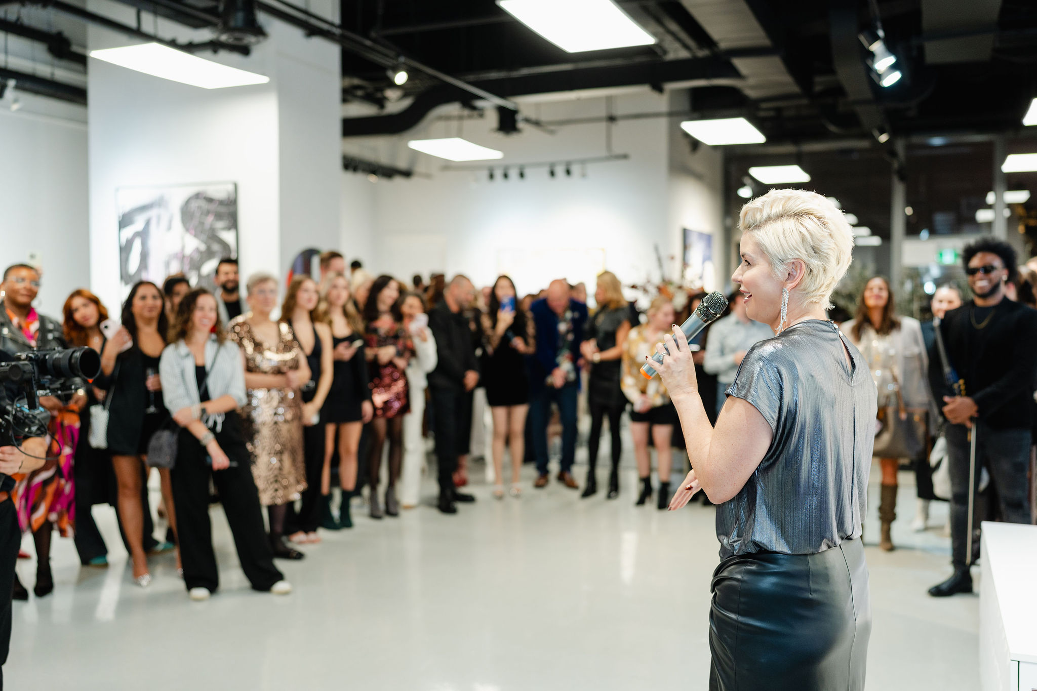 A person with short blonde hair speaks into a microphone at an indoor corporate photography event, with a group of attendees standing and listening.