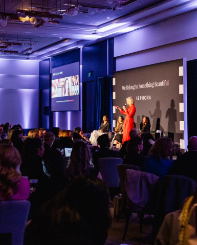 A speaker in a red outfit addresses an audience at a conference, captured in striking corporate photography. A large screen displays colorful images, and the backdrop reads "We Belong to Something Beautiful" with the Sephora logo.