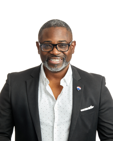 A person with glasses and a beard smiles in a corporate photography shoot, wearing a black blazer, white shirt, and lapel pin, posing against a white background.