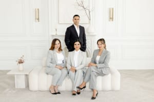 Four people in business attire pose in a white, minimalist room, epitomizing personal branding photography. Three are seated on a sofa, with one confidently standing behind them.