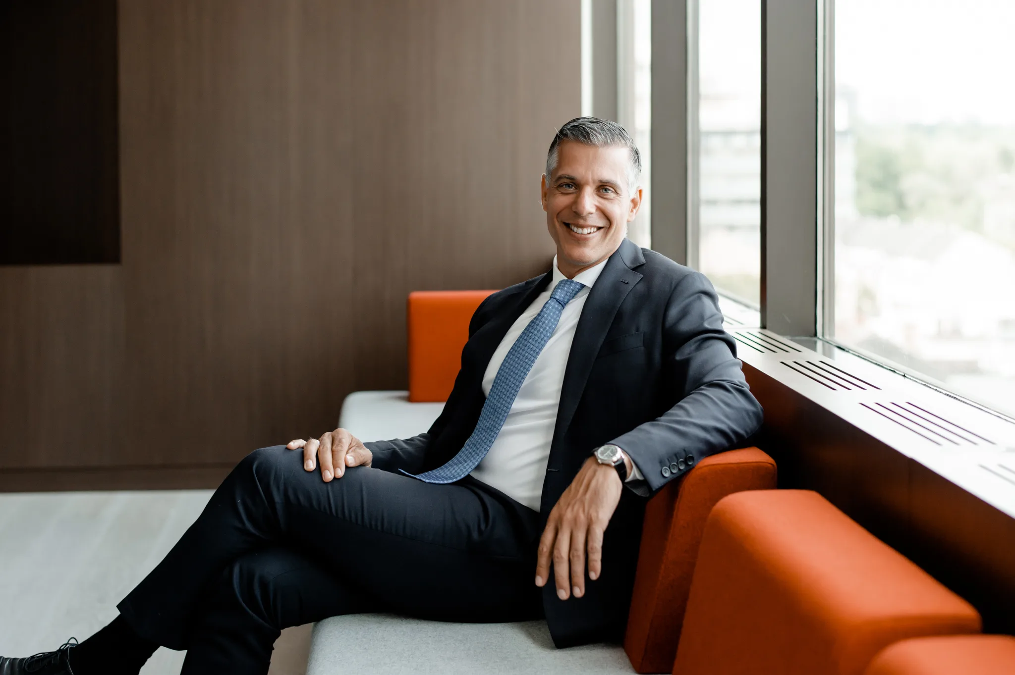 A man in a suit sits on a couch by a window, smiling. The room has modern decor, with wooden walls and orange accents.