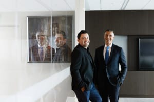 Two men in business attire stand smiling in an office; a painting of them is reflected in glass nearby.
