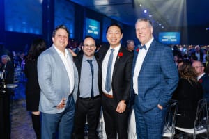 Four men in suits pose together at an indoor event, captured against a striking blue-lit background, showcasing the artistry of event videography.