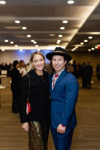 A woman and a man in formal attire pose together in a well-lit room, perfectly capturing the essence of event videography, with people bustling in the background.