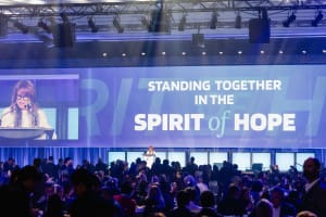 A speaker at a podium next to a large screen displaying "Standing Together in the Spirit of Hope" captivates the audience, as event videography captures every inspiring moment at the crowded gathering.