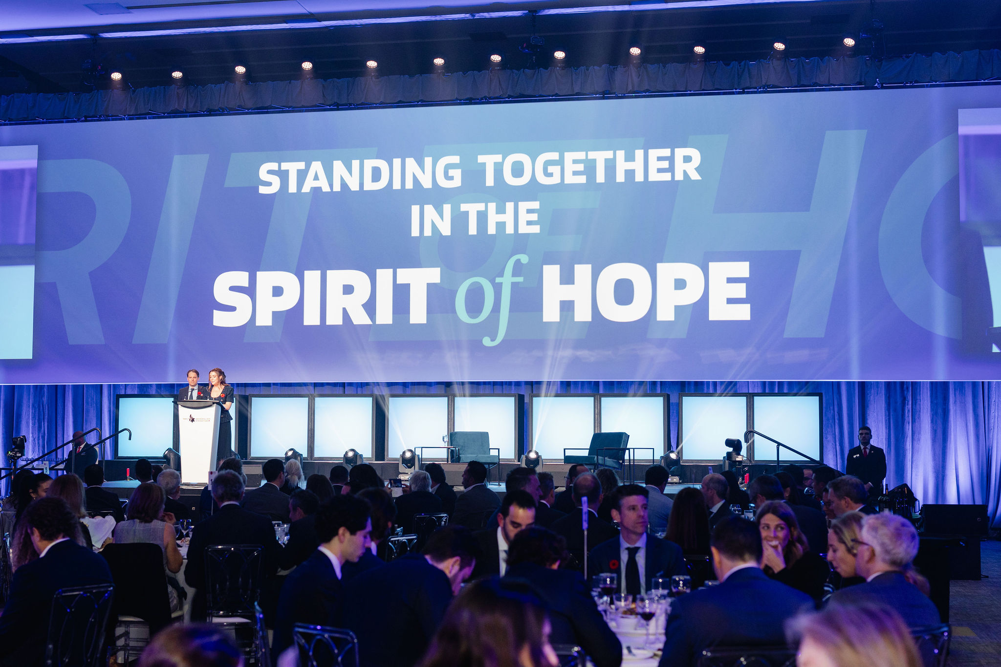 A large event with attendees seated at tables facing a stage adorned with a "Standing Together in the Spirit of Hope" banner, all captured expertly by event videography to preserve every inspiring moment.
