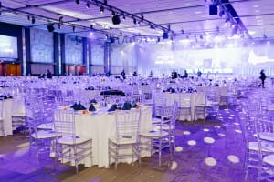 A large, elegantly lit banquet hall with round tables set for an event. Transparent chairs surround the tables, while purple lighting adds ambiance. An event videography team captures every detail, preserving the enchanting atmosphere for lasting memories.
