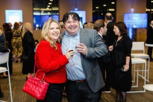 A man and a woman smiling and holding drinks at an indoor social event are captured in perfect harmony. The woman, donning a striking red jacket and matching handbag, radiates joy—ideal moments for event videography to preserve these cherished memories.