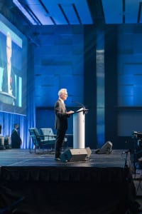 A person speaks at a podium on stage, as event videography captures their image projected onto a large screen in the conference setting.