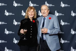 Two people smiling and holding drinks in front of a Simon Wiesenthal backdrop, wearing poppy pins, capture the spirit of the event. The moment is perfectly suited for event videography, allowing memories to be cherished forever.