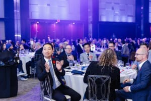 A large group of people sits at tables in a formal event setting, some clapping as the dim blue lighting sets the stage for expert event videography.