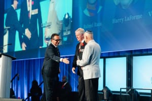 Three men on stage engage in a handshake and conversation during a formal event, captured through expert event videography, with a large screen displaying text in the background.
