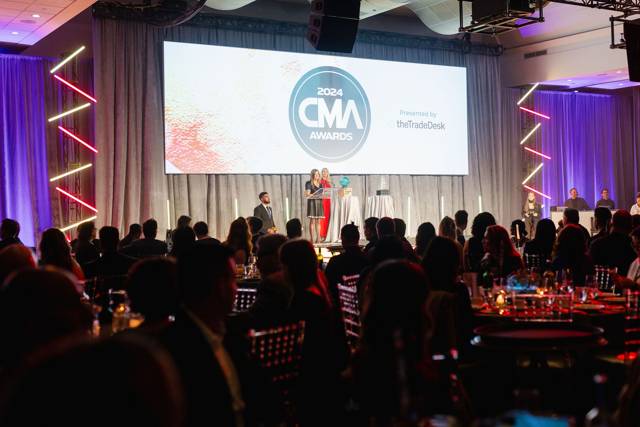 A person speaks at the podium during the 2024 CMA Awards ceremony, captured in stunning corporate photography. The audience is seated at tables in a large, elegantly decorated venue.