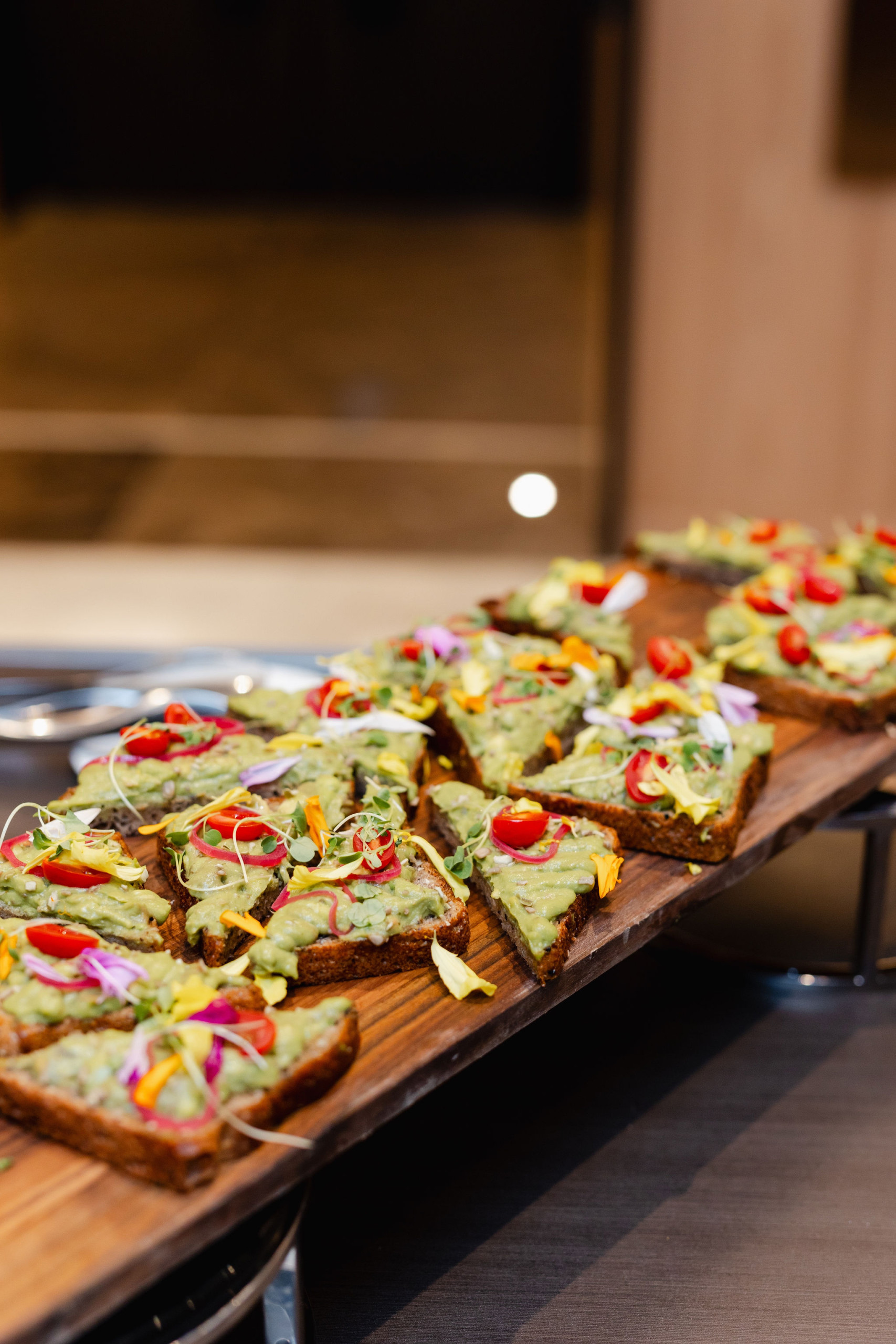 Slices of bread topped with avocado spread, cherry tomatoes, and edible flowers are artfully arranged on a wooden tray, showcasing exquisite brand photography.