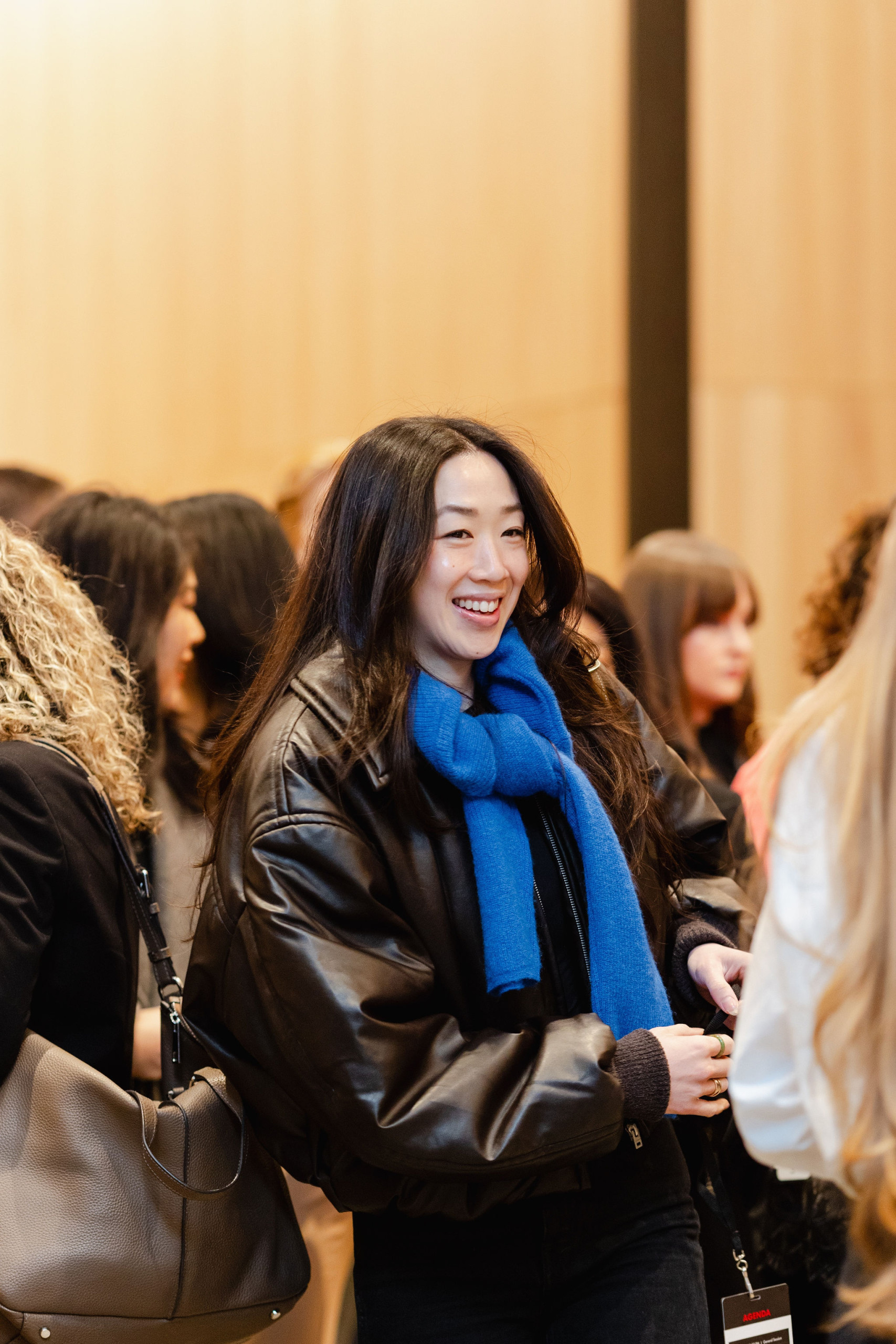In the bustling room, a person in a blue scarf and black jacket radiates joy as they smile, perfectly capturing the essence of brand photography.