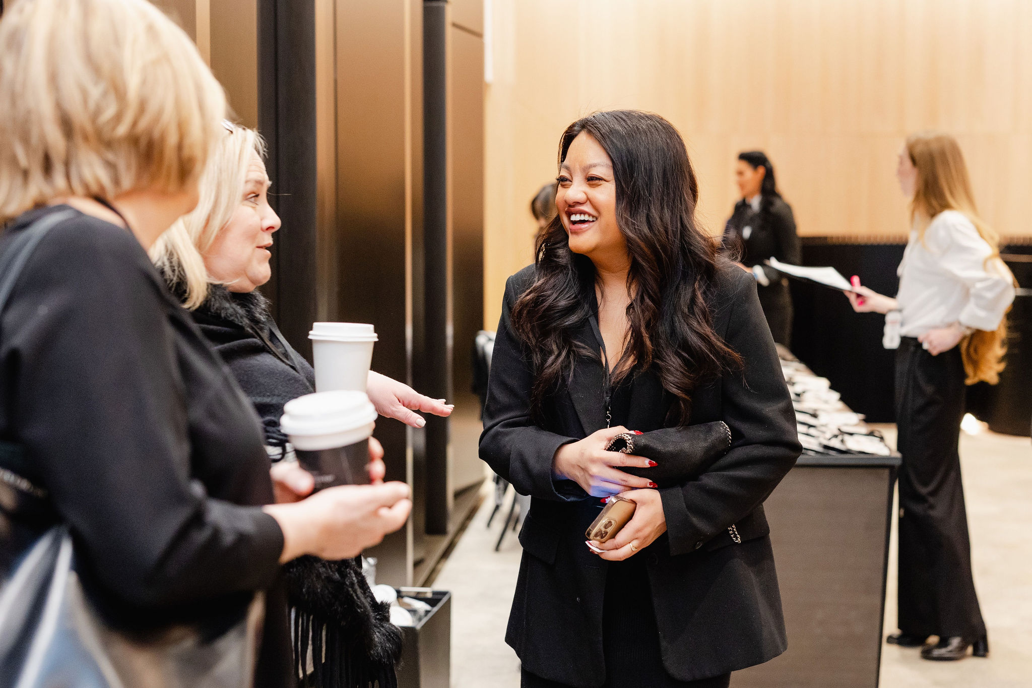 Three women in business attire engage in conversation, each holding a drink, as they are captured in an office setting perfect for brand photography.