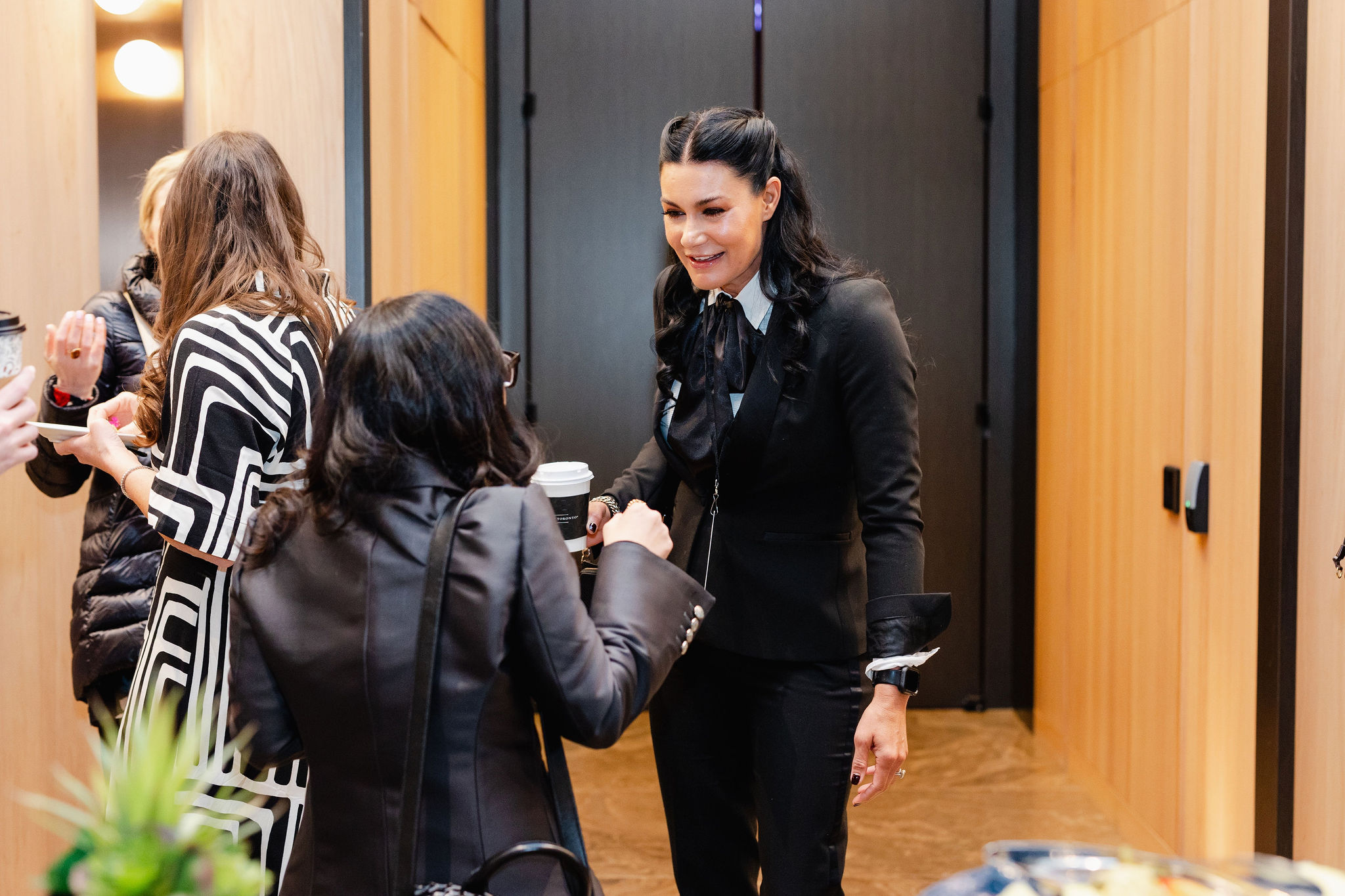 In a hallway, captured with the precision of brand photography, two women in formal attire warmly greet each other, one gracefully holding a coffee cup.