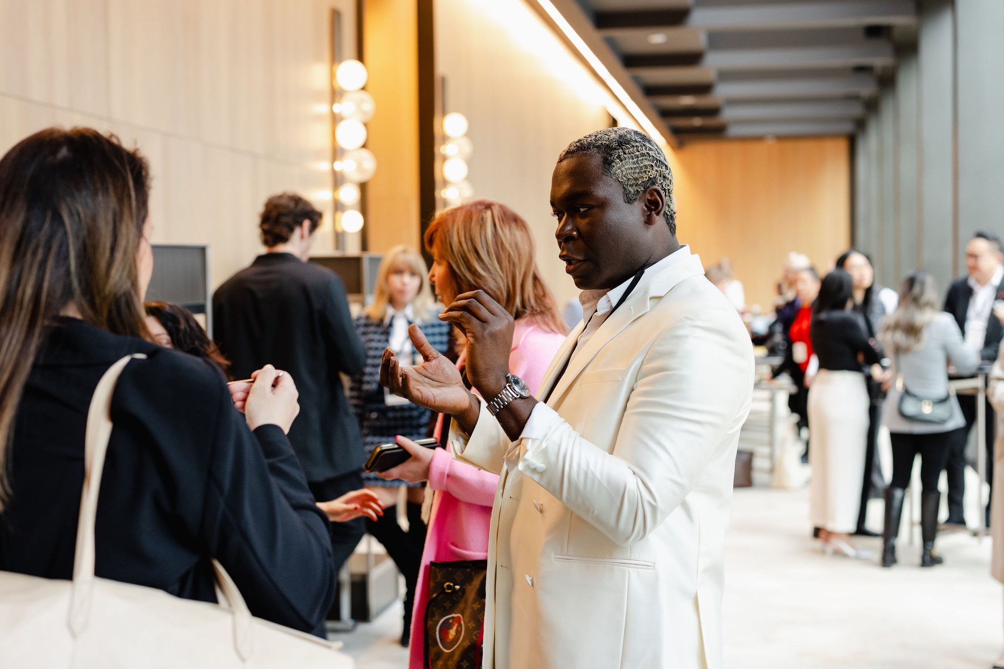 In a scene reminiscent of brand photography, a man in a white suit engages in conversation with a woman in a black jacket, their interaction poised amid the bustling backdrop of people moving through the lively room.