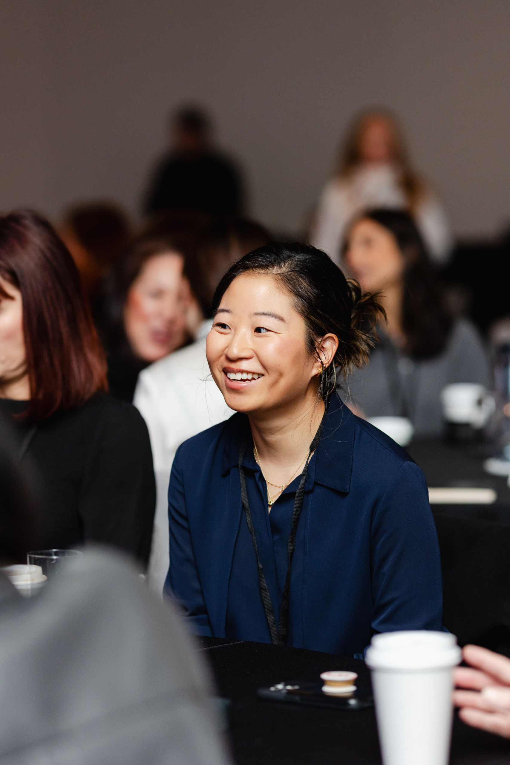 In a vibrant conference setting, a woman in a blue blouse sits at a table, smiling warmly. Her presence embodies the essence of brand photography, with several people around her adding to the dynamic atmosphere.