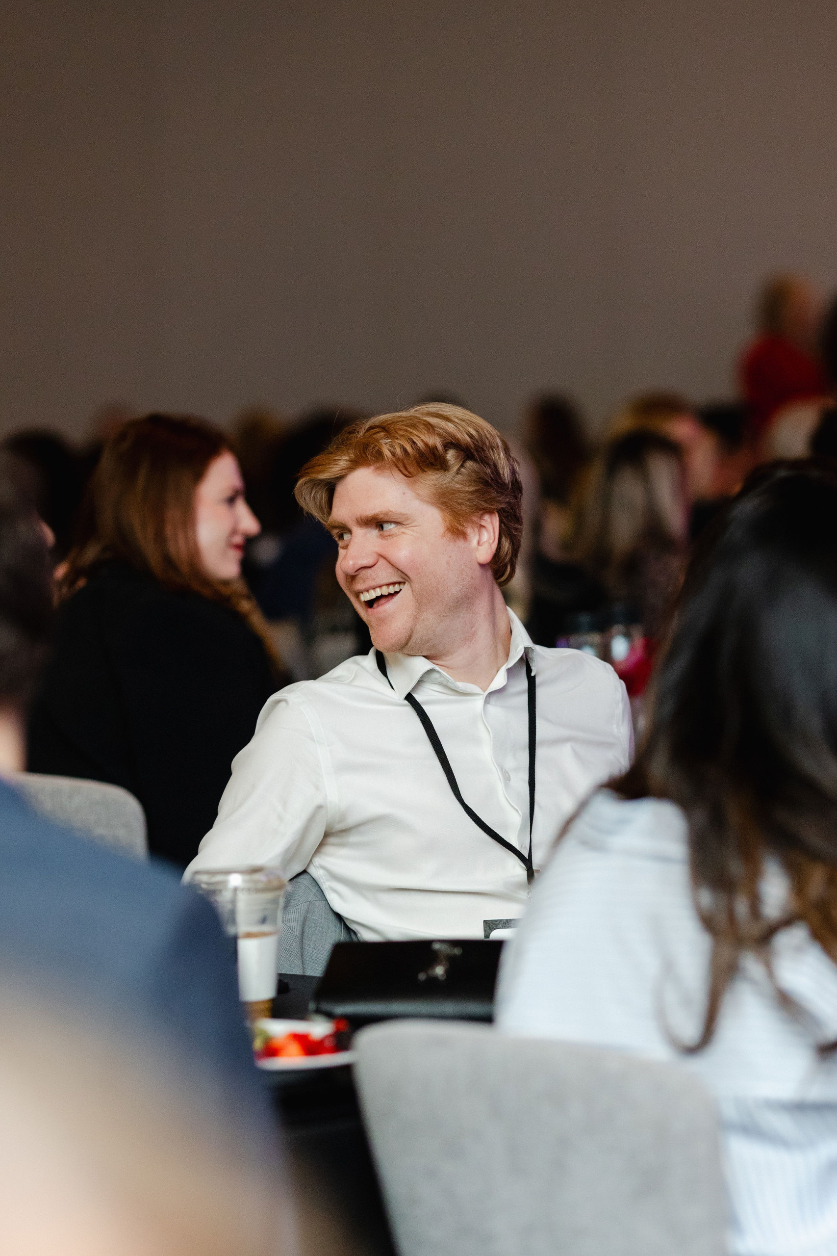A person in a white shirt is smiling while seated among others in a conference setting, capturing the essence of authentic brand photography.