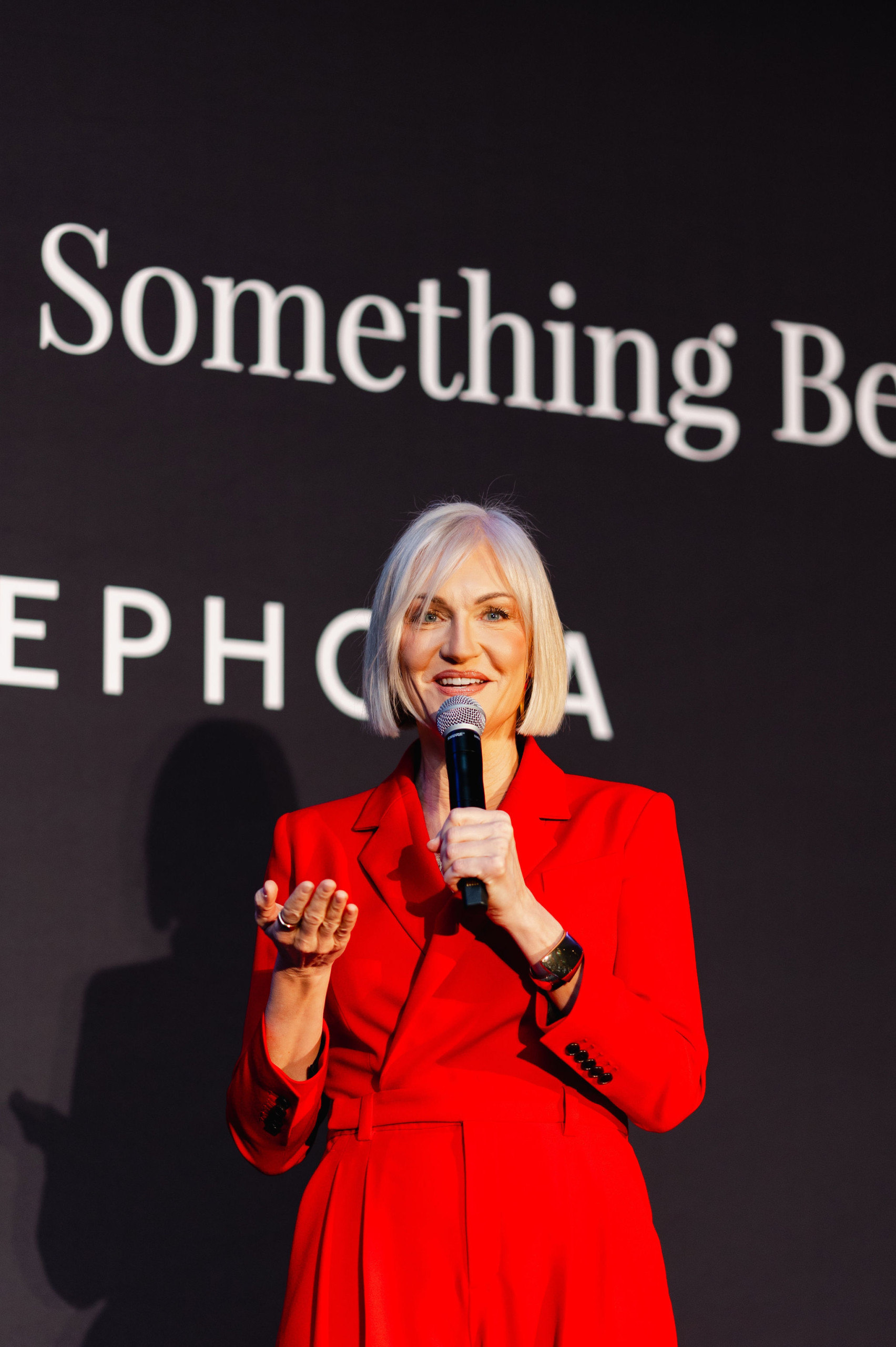 A person in a red outfit speaks into a microphone on stage, with a black background showcasing partial brand photography text.
