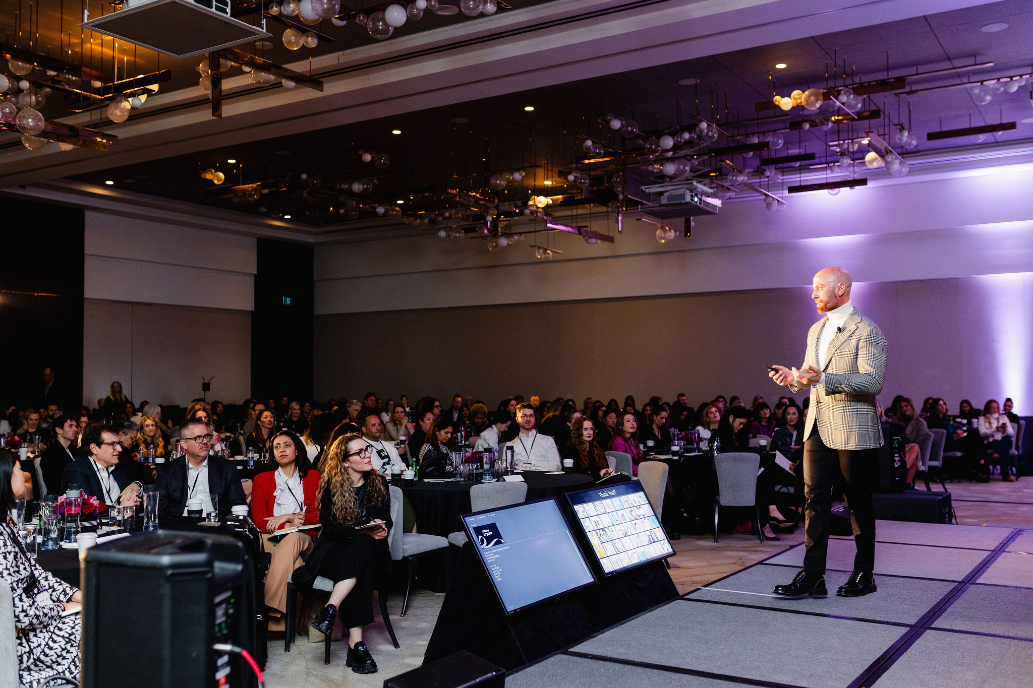 A speaker standing on stage addresses an audience seated at round tables in a conference room, capturing the essence of brand photography through carefully curated visuals that enhance the event's atmosphere.