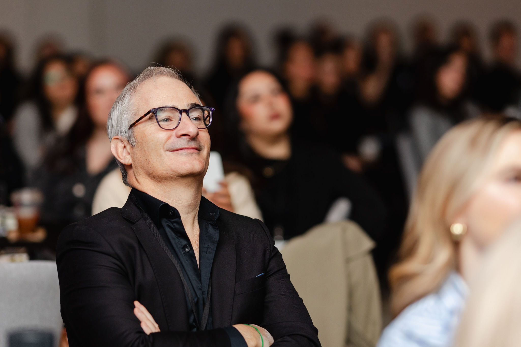 A man in glasses and a black suit sits with arms crossed, smiling confidently amidst the blurred backdrop of an audience, embodying brand photography at its finest.