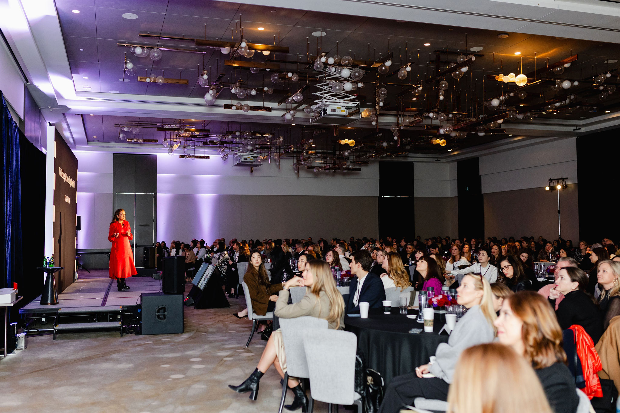 A speaker in red engages a seated audience in a conference room with a modern ceiling design, capturing the essence of brand photography.