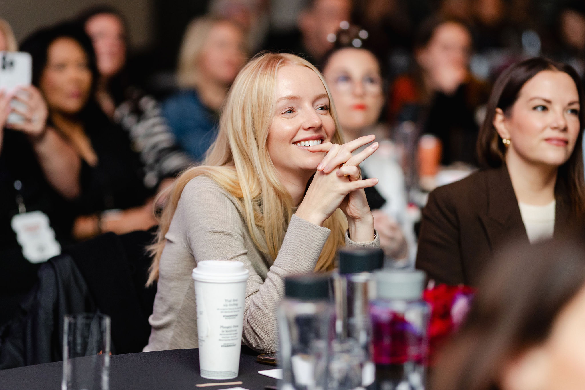 A group of people sitting at a conference, with a blonde woman smiling in the foreground, capturing the essence of brand photography. A coffee cup and water bottles are on the table.