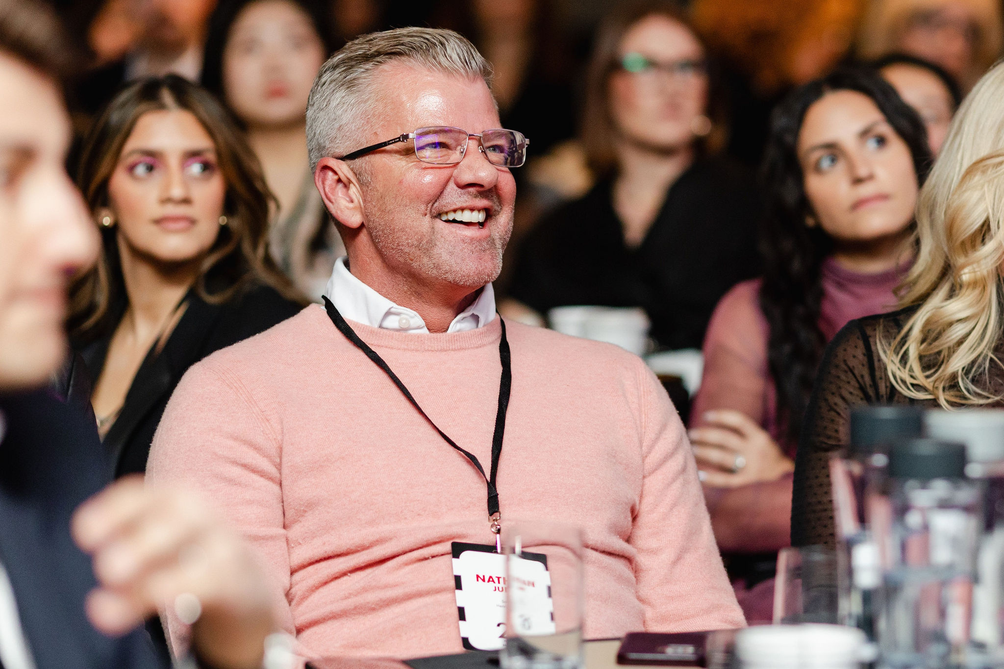 A smiling man in a pink sweater and glasses sits among a group of people, capturing the essence of brand photography. He has a conference name tag around his neck.