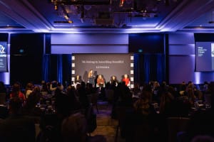 A panel of five people sits on a stage with the backdrop "We Belong to Something Beautiful" at a conference, capturing the essence of brand photography. The audience is seated in the foreground, perfectly framing this moment of inspiration and connection.