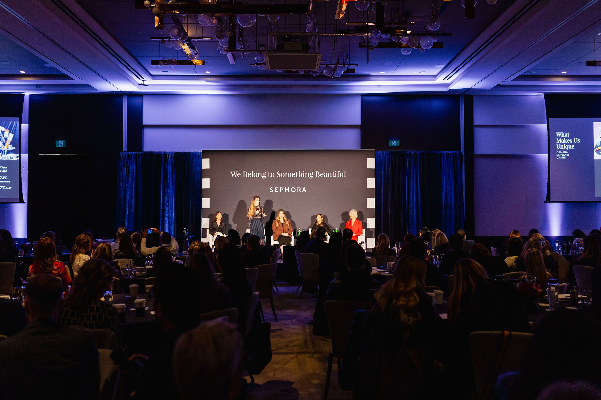 A panel of five people sits on a stage with the backdrop "We Belong to Something Beautiful" at a conference, capturing the essence of brand photography. The audience is seated in the foreground, perfectly framing this moment of inspiration and connection.