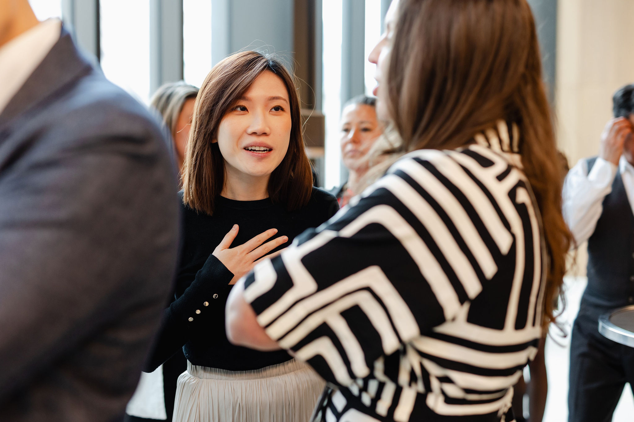 At a social event, two people converse while artfully captured in brand photography. A woman with brown hair and a black sweater places her hand on her chest as she speaks, with others partially visible in the background.