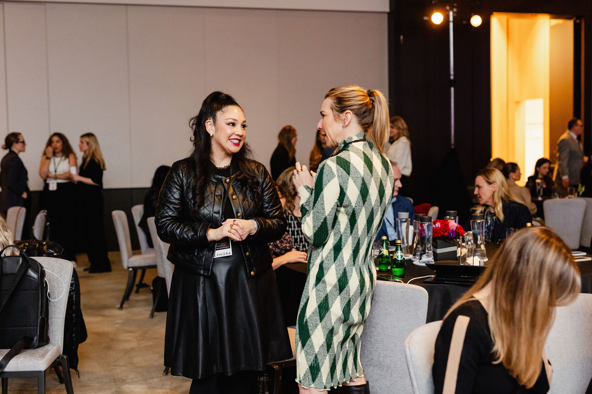 Two women are standing and conversing at an indoor event, showcasing vibrant brand photography. More people mingle in the background amid several tables and chairs, creating a lively atmosphere.