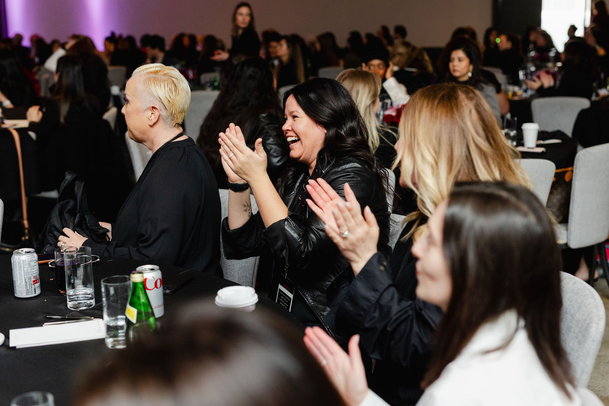 Audience members sit at a table, clapping and smiling during an indoor event, with drinks and brand photography capturing the joyous moment.