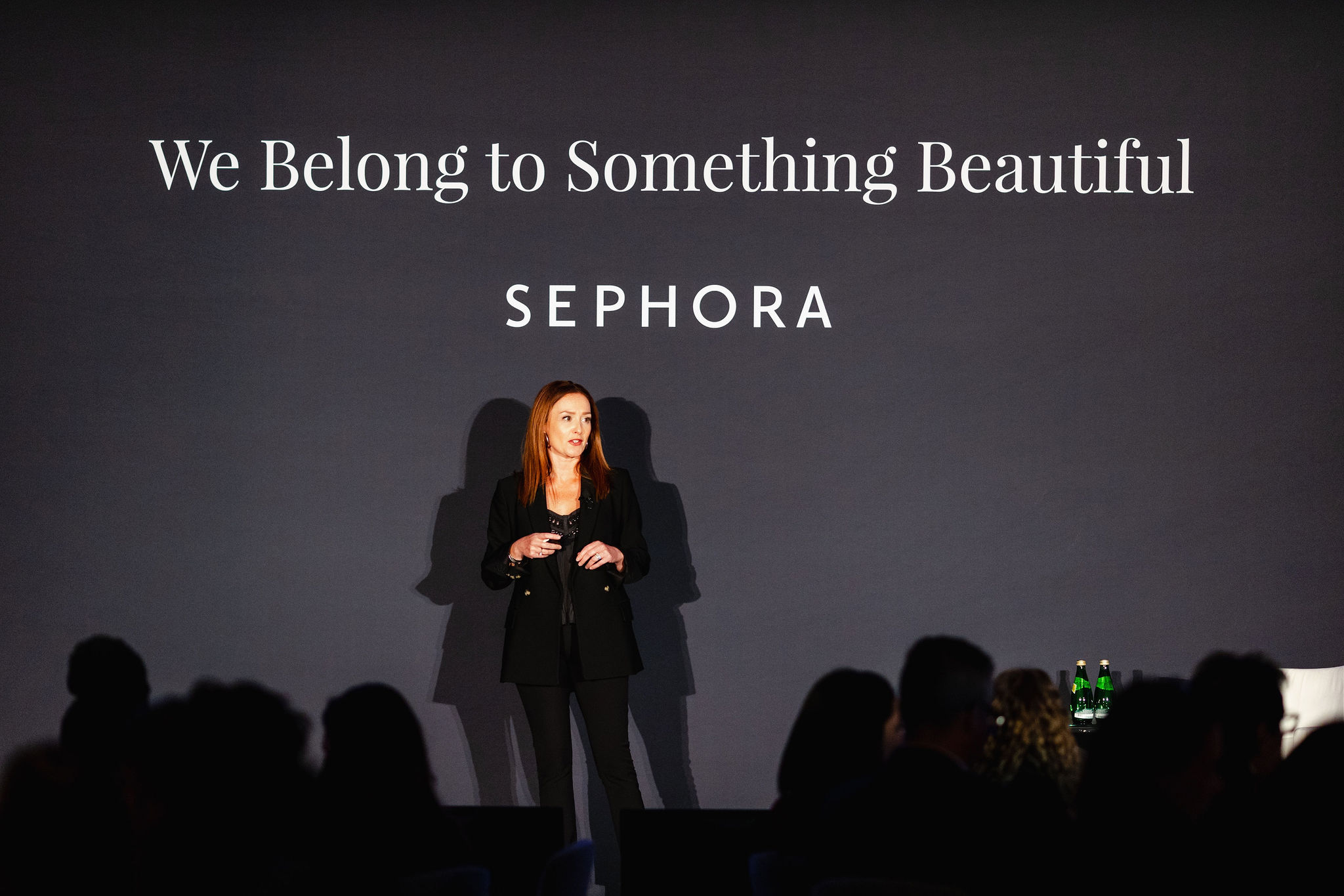 A woman in a black outfit stands on stage, embodying the essence of brand photography, against a gray backdrop with the Sephora slogan, "We Belong to Something Beautiful," written in white.