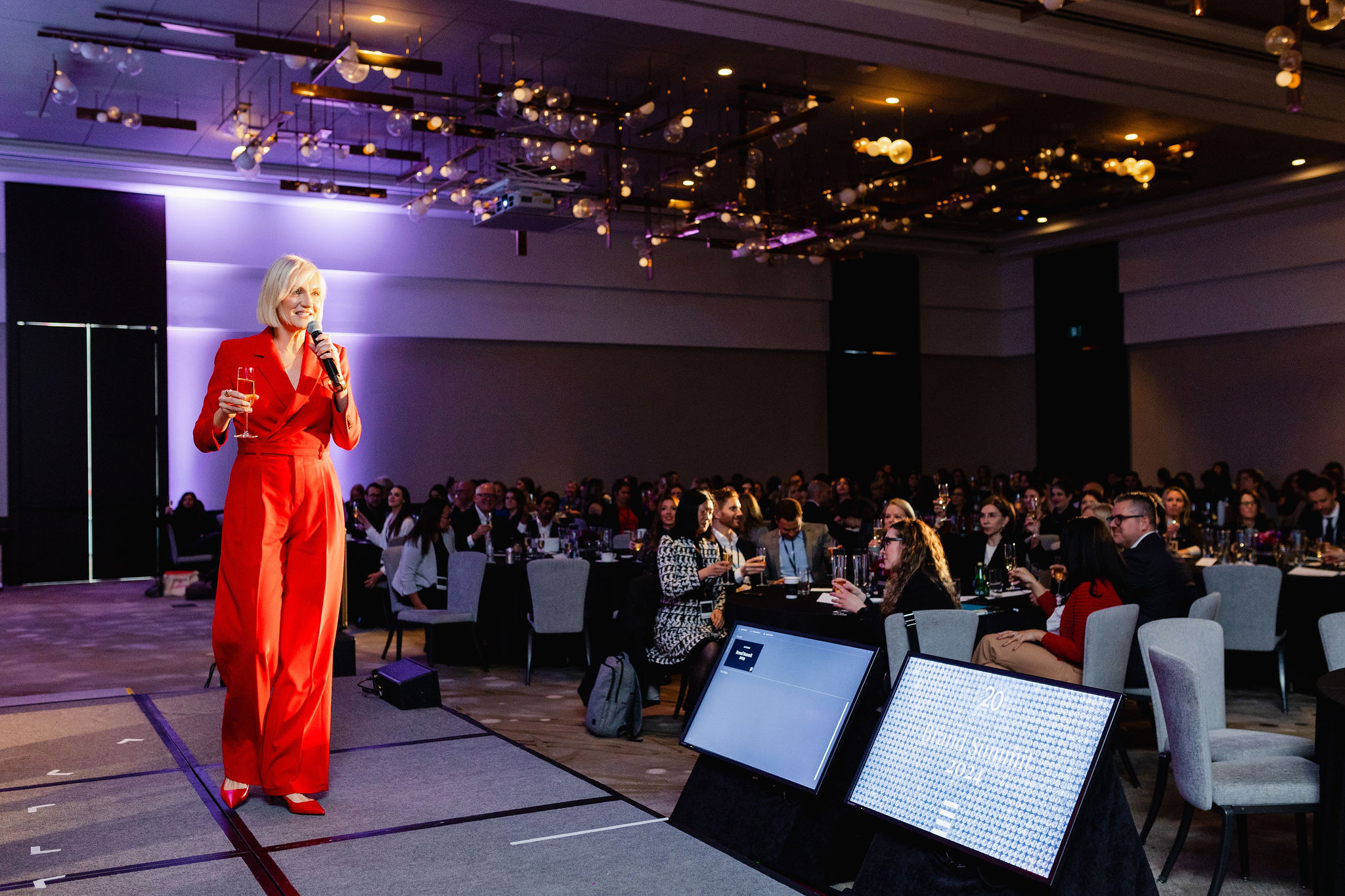 A person in a red outfit captivates the audience, speaking into a microphone on stage at an event. The scene, perfect for brand photography, features attentive seated audience members and multiple screens enhancing the dynamic atmosphere.