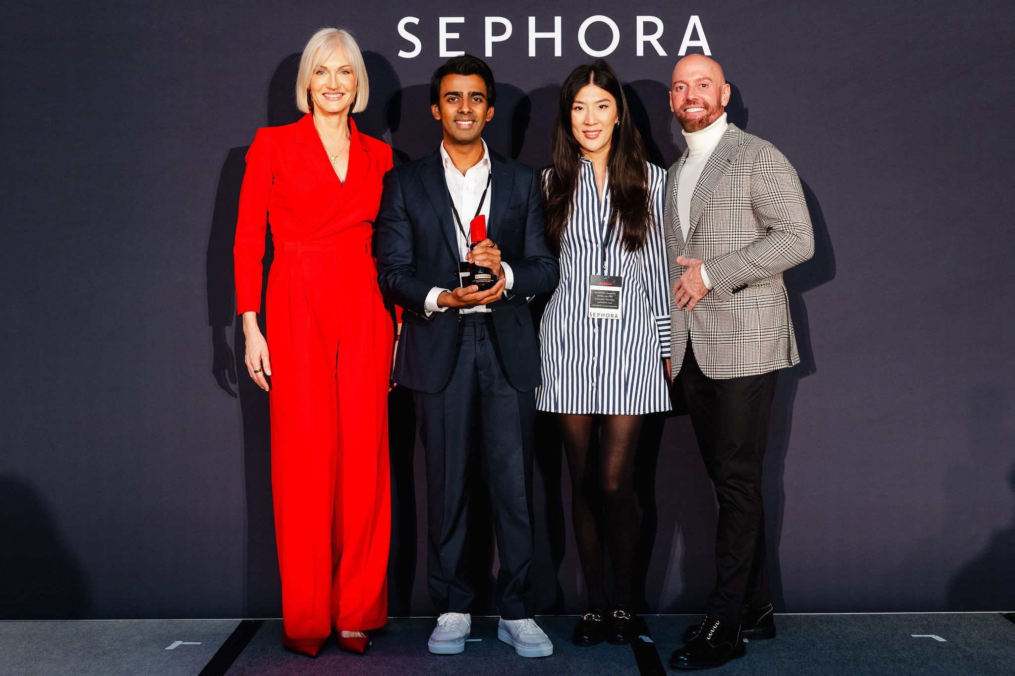 Four people stand on a stage in front of a Sephora backdrop, exuding elegance. One holds a trophy, embodying the spirit of brand photography. They are impeccably dressed in formal attire: a striking red suit, classy navy suit, chic striped dress, and stylish plaid blazer.