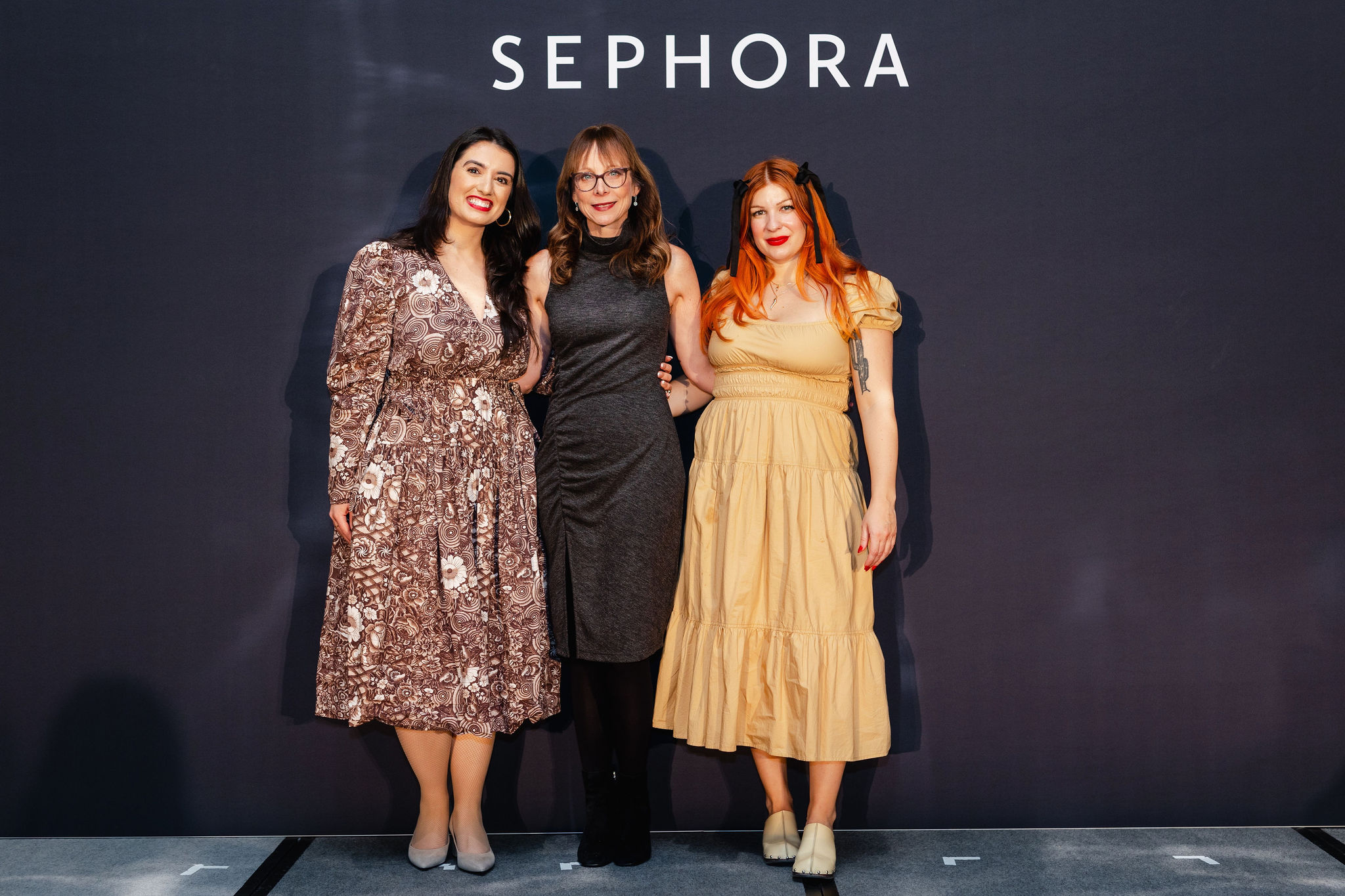 Three women pose confidently in front of a black backdrop adorned with "Sephora," showcasing brand photography at its finest. Dressed in floral, grey, and beige dresses, their smiles light up the scene.