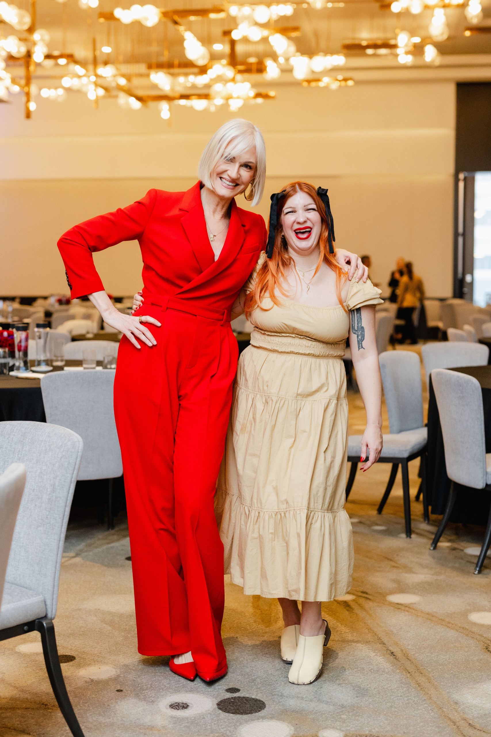 Two people are smiling and posing together in a room adorned with tables and chairs. One dons a red suit, the other a beige dress. This moment captures the essence of brand photography, highlighting style and personality in perfect harmony.