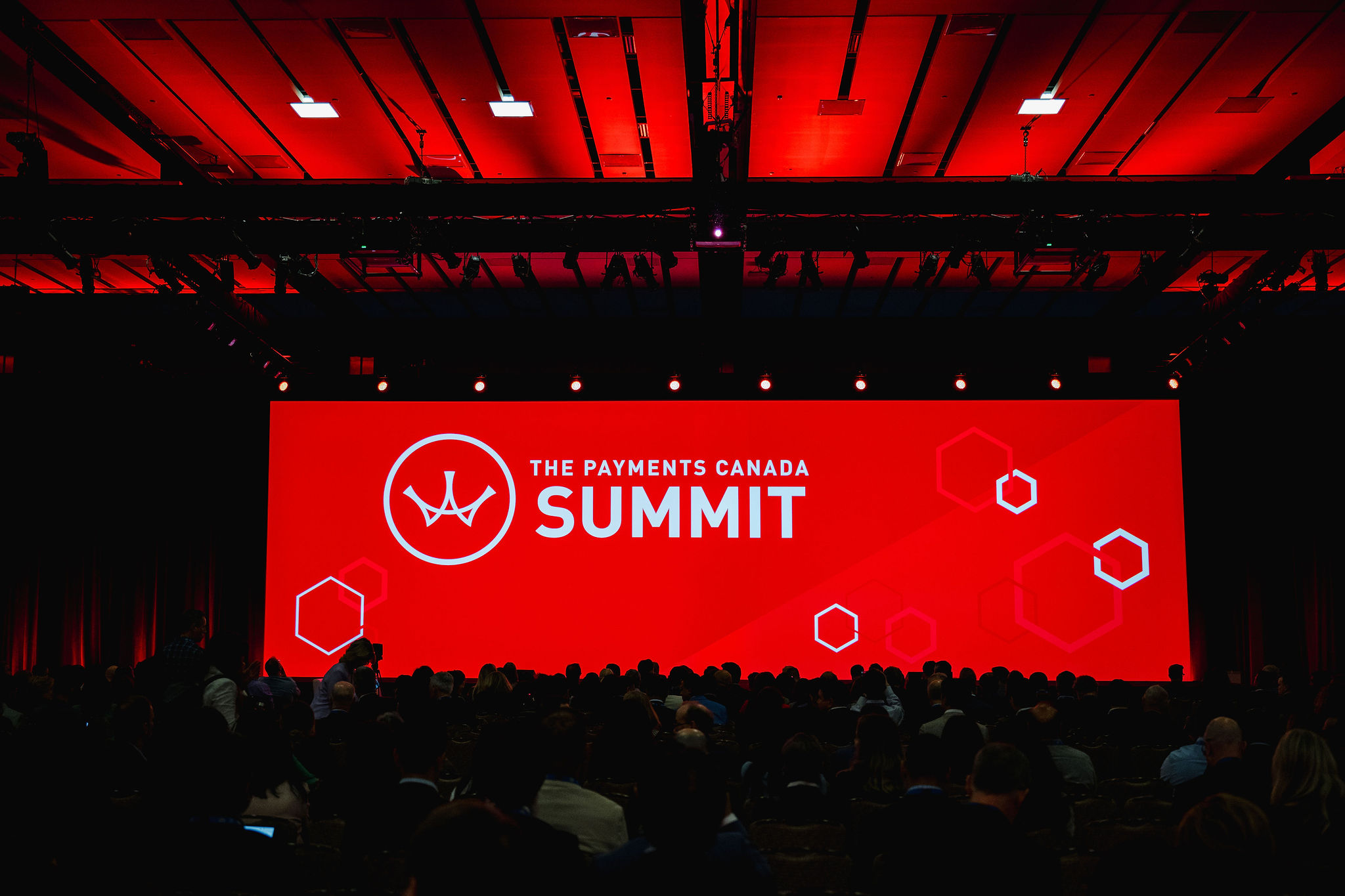 A large screen displays "The Payments Canada Summit" logo in a dimly lit room, capturing the essence of event photography with an attentive audience seated below.